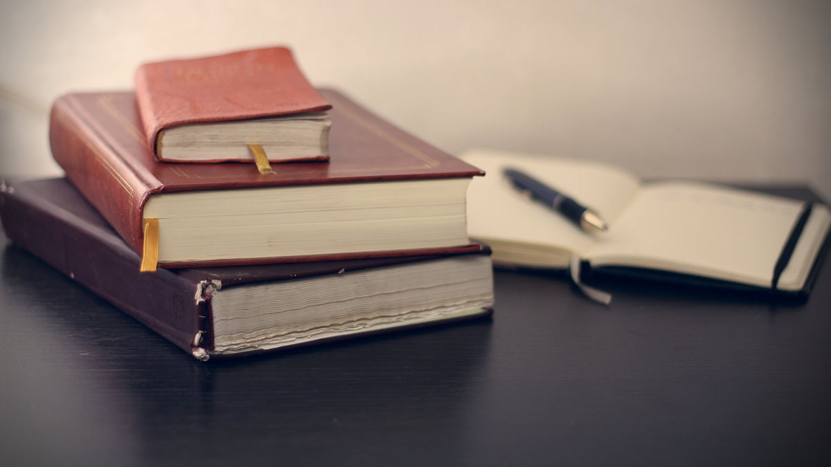 Aufeinanderliegende Bücher neben einem offenen Notizbuch mit Stift drauf